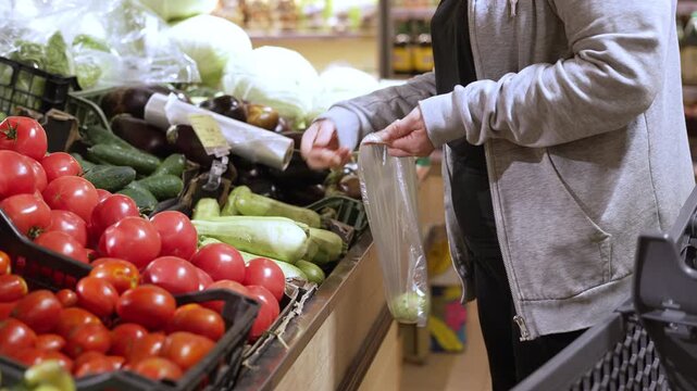 Customer selecting zucchini and cucumbers at supermarket produce counter. Fresh vegetables placed into plastic bag for grocery shopping and healthy food concept