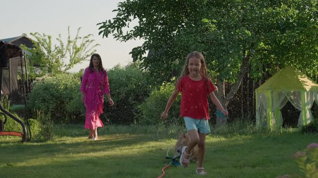 Family Outdoor Activity, Woman And Child Enjoy Sunny Day, Playful Dog Chasing Fountain In Garden Scene, Cheerful Family Spending Time Outdoors With Their Energetic Dog And Blooming Plants