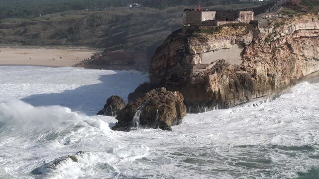 Cinematic aerial drone establishing shot of big waves breaking on rocks and cliffs on a day with giant waves in Nazar&eacute;, Portugal, Europe. Farol da Nazare lighthouse and wind turbines in the background