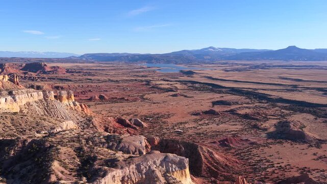Drone footage drifts above rugged red rock cliffs into sweeping desert plains, revealing layered badlands, scattered shrubs, winding natural patterns, and distant mesas under clear skies.
