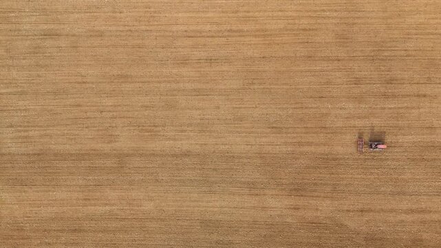 Top down shows a small crawled tractor working a large, golden‑brown field in rural Tuscany, Italy, smoothing and leveling the soil in gently rolling farmland bordered by a strip of green vegetation.