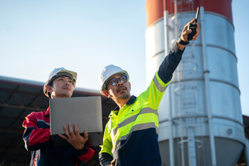 Two engineers wearing safety helmets discussing site conditions while reviewing data on a laptop, standing in front of an industrial silo during daylight inspection.