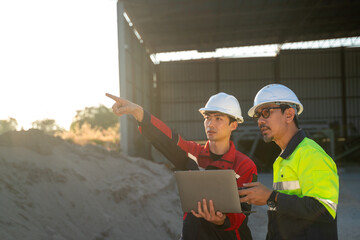 Engineers standing in open industrial area, analyzing project information on a laptop with silo and factory buildings in the background.