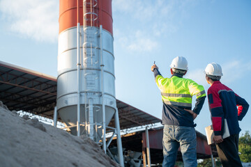 Rear view of two engineers observing a tall industrial silo, one pointing upward while holding a communication device, emphasizing inspection and leadership.