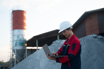 An operations manager reviewing raw material logistics on a portable computer, set against a backdrop of aggregate piles and factory structures.