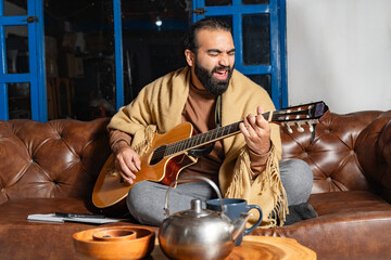 Person singing and playing guitar cross-legged on leather couch with tea and notebook
