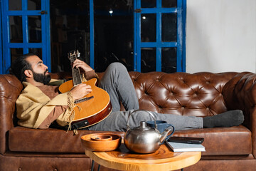 Person reclining with guitar and tea in cozy indoor setting with notebook and phone