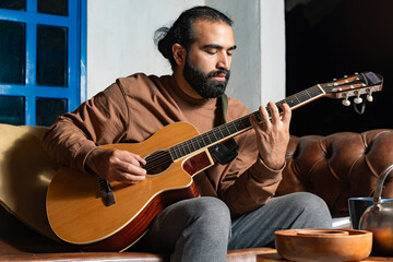 Person playing acoustic guitar on leather couch with teapot and bowls nearby