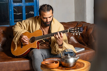 Person playing guitar with mug and teapot on table in intimate indoor setting