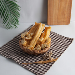 A wicker basket filled with various types of breadsticks and pastries sits on a black and white checkered cloth, with a wooden cutting board and a leafy branch in the background, on a white