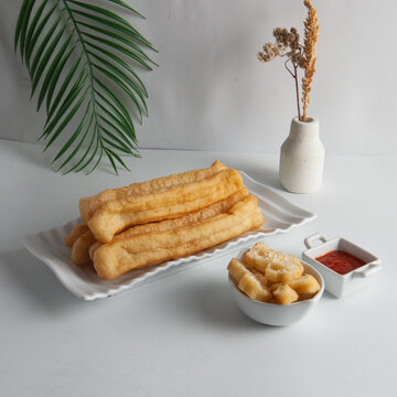 A plate of crispy fried churros with a bowl of dipping sauce and a side of cakwe on a white table with a minimalist background and warm tones.
