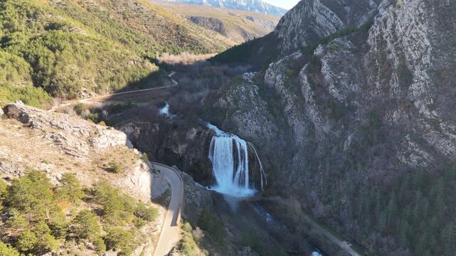 Drone flying backward from Krčić Waterfall, revealing an ever-widening panorama of the Krka River, Napoleon road, curving gorge, and massive karst mountains near Knin, Croatia.