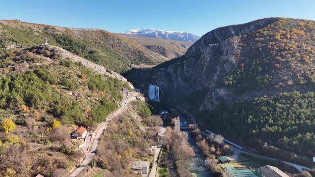 Wide aerial panorama as the drone approaches Krčić Waterfall near Knin. Snow-capped Mount Dinara in the distance, shaded river gorge, and green Krka River below on a sunny day.