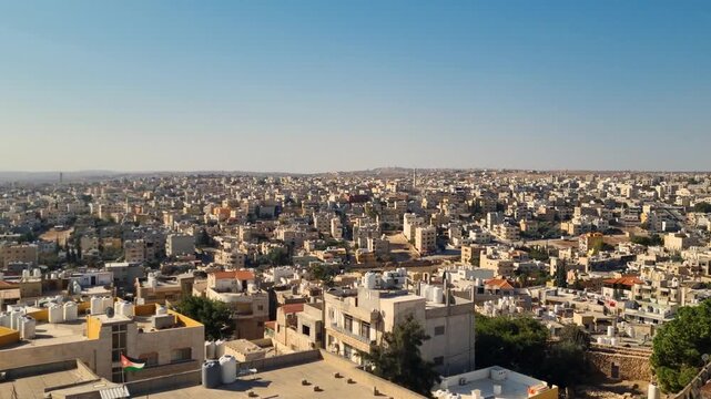 Wide skyline view of Madaba, Jordan, under the midday sun, with light washing over rooftops as the city spreads across rolling hills toward a distant horizon