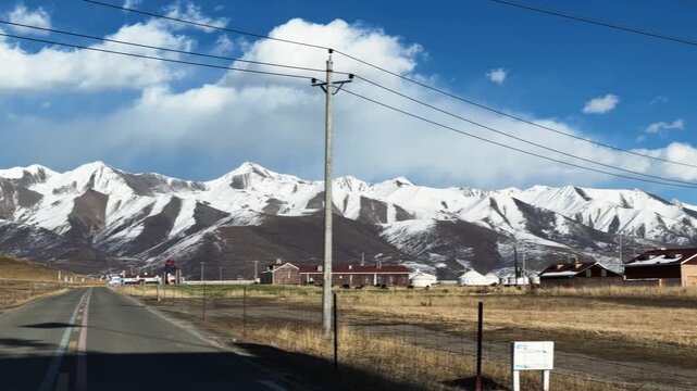 Qilian Grassland, Xining, Qinghai Province, China - A Straight Country Road Stretches Toward Snow-covered Mountains, Passing Quiet Grasslands and Small Buildings Beneath a Blue Sky - Pan Right Shot
