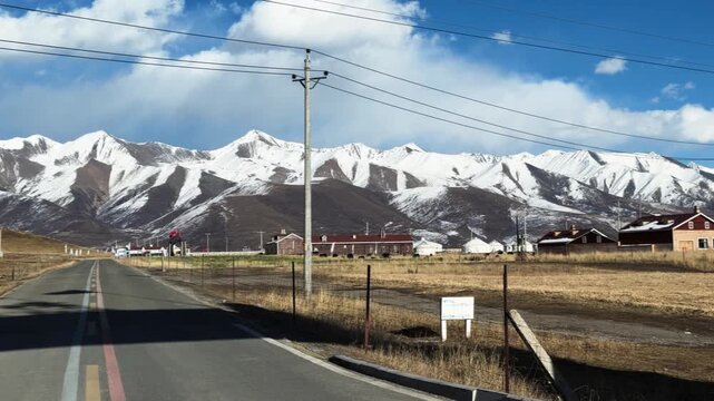 Qilian Grassland, Xining, Qinghai Province, China - A Quiet Rural Road Leads Toward Snow-capped Mountains Under a Wide Blue Sky, with Scattered Buildings Across the Open Grassland - Pan Right Shot
