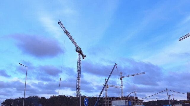 Cranes lift materials at a building site. Workers are busy with construction tasks. The sun is shining and clouds are scattered in the sky.