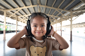 Happy child with headphones, smiling girl at train station, casual face portrait, joyful travel vibe, candid outdoor scene, soft natural light, warm tone mood