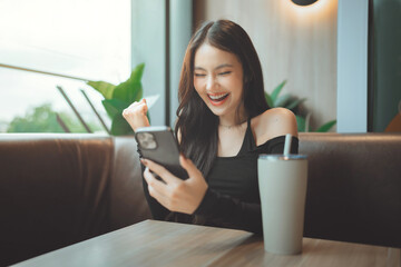 Happy young asian woman cheering with fist pump while looking at smartphone in cafe for good news business success or winning online concept