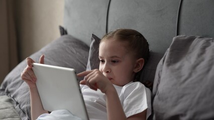 child playing tablet, little girl playing tablet, comfort zone for kids, girl wearing white shirt, comfortable home environment, gray sheets and calm colors, relaxed girl watching videos tablet in bed