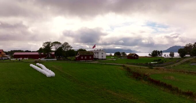 Aerial drone view of Kvitnes G&aring;rd, a historic farm and restaurant in Vester&aring;len, Norway. Traditional red and white buildings with sod roofs sit amidst green fields near the fjord under a cloudy sky