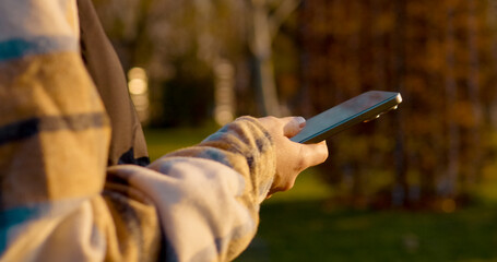 Using smartphone while walking in the park bench during golden autumn afternoon, woman browsing digital content in soft sunlight