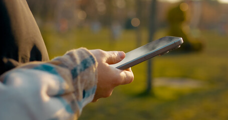 Woman millennial networking on smartphone while relaxing in urban park during golden hour sunset, casually enjoying digital connection amid natural landscape