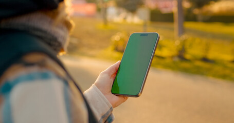 Woman navigating smartphone with green screen while walking through sunlit park, embodying modern...