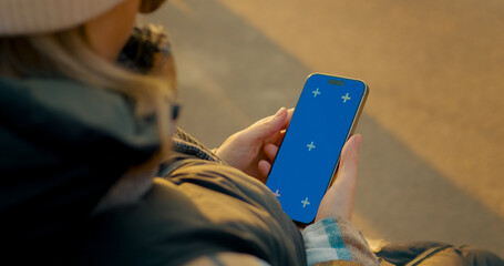 Female relaxing on outdoor bench during winter, holding smartphone with blue chroma key screen for...
