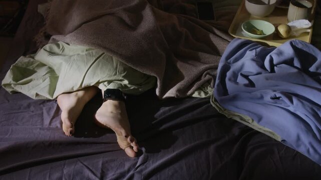 High angle shot of unrecognizable woman lying in bed, covered by blankets, bare feet with house arrest ankle bracelet sticking out