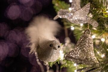Close-up of silver Christmas bird ornament with white feather details, hanging on a decorated tree with warm white LED lights and soft bokeh background.