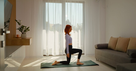 A young redhead performs yoga in a well-lit living room with large windows. The morning light enhances her mindful exercise routine, promoting a healthy lifestyle.