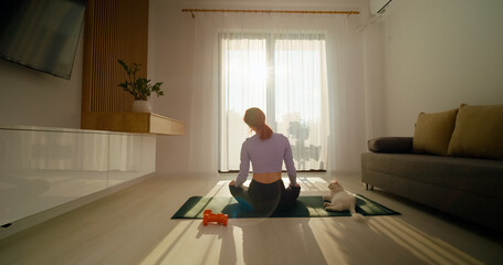A redhead woman engages in yoga and meditation in her bright living room, creating a peaceful atmosphere as her cat relaxes nearby in the morning sunlight.