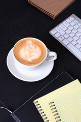 A cup of coffee on a black background. A notebook for writing and a computer keyboard on the table. The concept of a business breakfast or coffee break. Food and drinks.