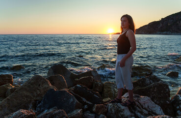 A girl poses and relaxes by the sea, on the rocks, a panoramic view of the coast and mountains at sunset, sunlight creates backlighting and silhouettes