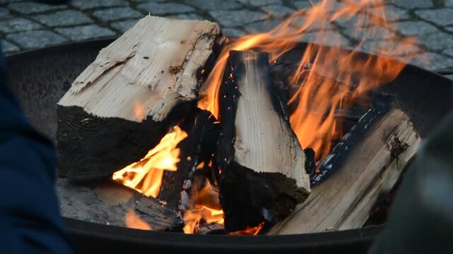 Close-up of split wood logs burning with bright orange flames in a fire bowl against a blurred cobblestone background, creating a cozy warmth