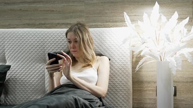 A girl is relaxing in her bedroom at dawn. She is nestled in bed under a blanket, and she scrolls on her phone. White plumes adorn her bedroom.