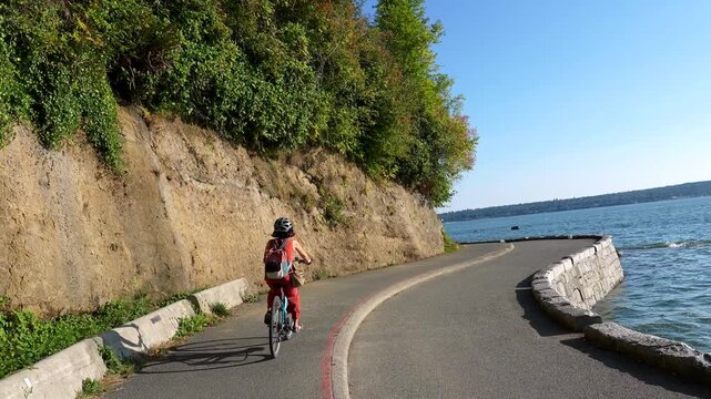 Woman cycling along scenic stanley park seawall in vancouver, enjoying ocean views, lush greenery, and coastal landscape during summer recreational bike ride