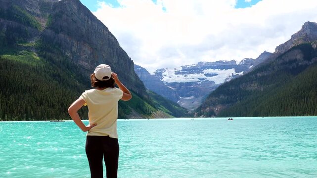 Female tourist wearing baseball cap standing at edge of turquoise lake louise, gazing at victoria glacier and surrounding mountains in banff national park, alberta, canada