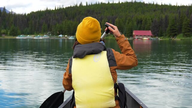 Tourist wearing a yellow beanie and a yellow life jacket paddles a canoe on maligne lake, surrounded by the beautiful scenery of jasper national park, with a red cabin visible on the shore
