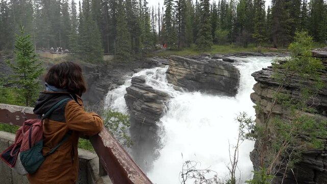Young female tourist wearing a brown jacket and backpack leaning on a wooden railing, contemplating athabasca falls in jasper national park, canada