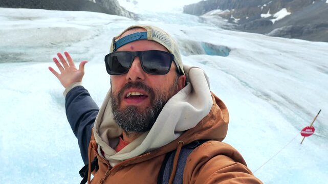 Happy tourist wearing sunglasses and a brown jacket showing with his hands athabasca glacier in columbia icefield, jasper national park, alberta, canada