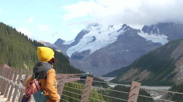 Female traveler wearing yellow beanie and backpack standing on glacier skywalk, admiring panoramic views of athabasca glacier in jasper national park, alberta, canada