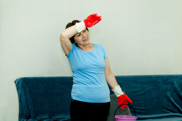 tired elderly woman in protective red rubber gloves holds bucket, wipes sweat from her forehead, resting after intensive house cleaning