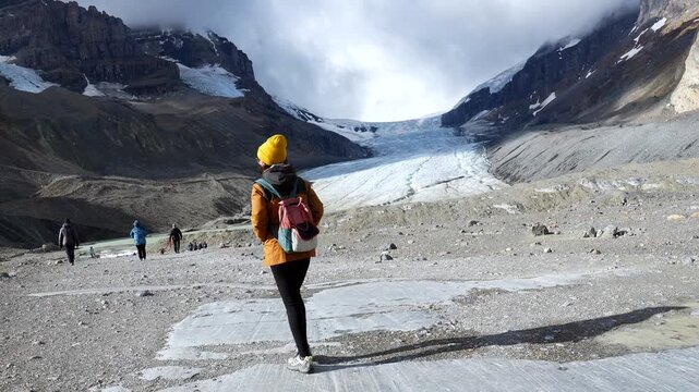 Young woman tourist with yellow hat and backpack walking on a path towards athabasca glacier in jasper national park, alberta, canada, in the columbia icefield area of the canadian rockies