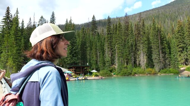 Female tourist wearing a cap and backpack admires the stunning turquoise waters of emerald lake, surrounded by a dense coniferous forest and mountain range in banff national park, canada