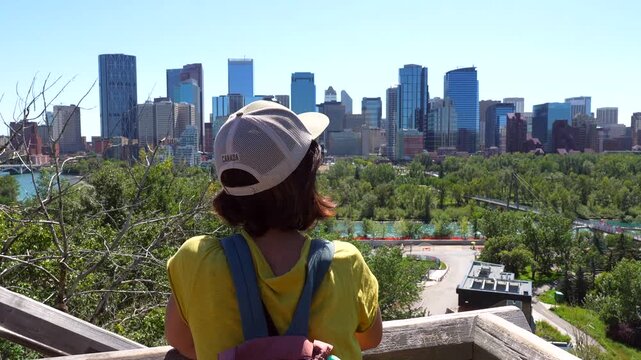 Female tourist wearing a baseball cap and backpack observes the cityscape of calgary, alberta, from a wooden viewpoint, showcasing urban skyline, lush greenery, and a clear blue sky