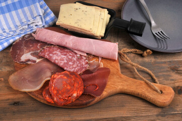 Assortiment de charcuteries sur une planche &agrave; d&eacute;couper en bois avec du fromage &agrave; raclette tranch&eacute; dans un po&ecirc;lon