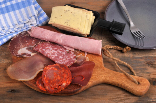 Assortiment de charcuteries sur une planche &agrave; d&eacute;couper en bois avec du fromage &agrave; raclette tranch&eacute; dans un po&ecirc;lon