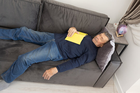 Elderly man lying on sofa indoors holding yellow book on chest relaxed posture home interior calm atmosphere.